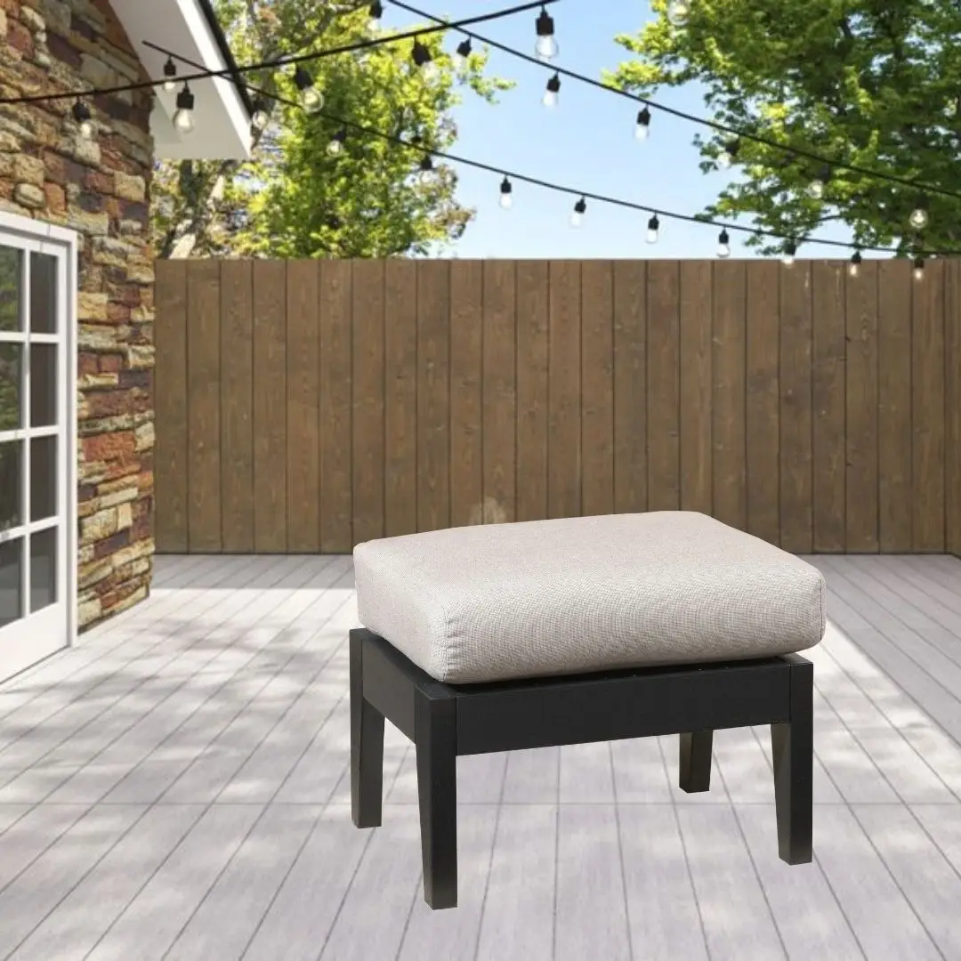 Outdoor patio scene with a black wooden ottoman topped by a light beige cushion on a gray deck, string lights overhead and a wooden fence in the background.