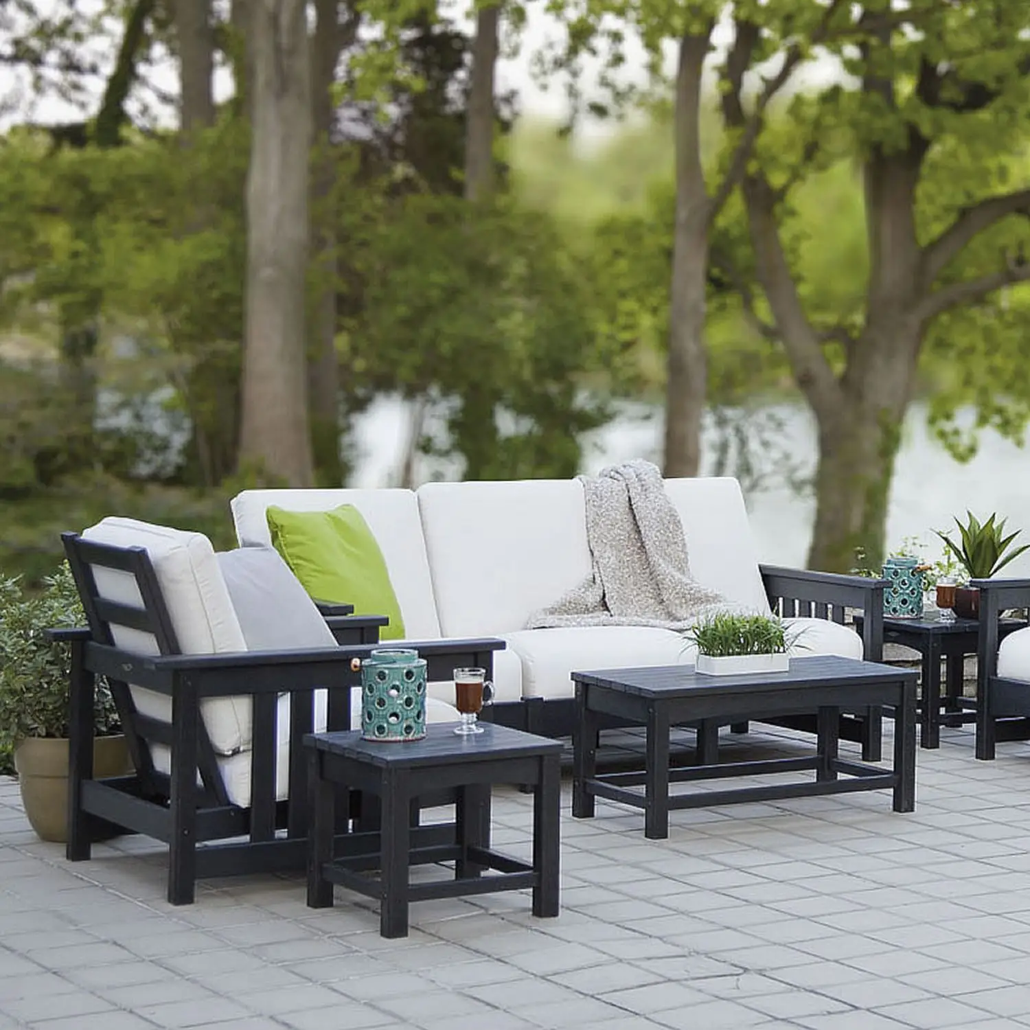 Outdoor patio seating set with white cushions and black frames on a tiled deck overlooking a lake and green trees, with a small table and potted plants nearby.